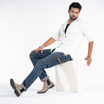 Man sitting on a white stool wearing a white shirt, blue jeans, and brown boots on a white background